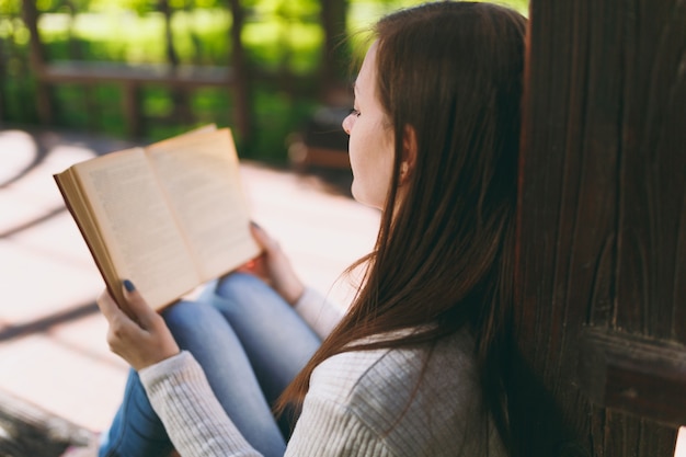 Portrait of calm peaceful young woman wearing light casual clothes relaxing, reading book. Female resting in city park in street outdoors on spring nature. Lifestyle concept. Back view. Copy space.