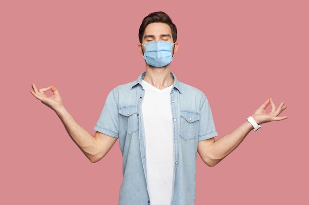 Photo portrait of calm relaxed young man with surgical medical mask in blue shirt standing with raised arms and doing yoga meditating exercise. indoor studio shot, isolated on pink background.