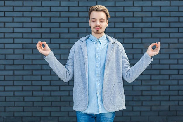 Portrait of calm serious handsome young blonde man in casual style standing in yoga pose, closed eyes and relaxed doing meditation. indoor studio shot on brick wall background.