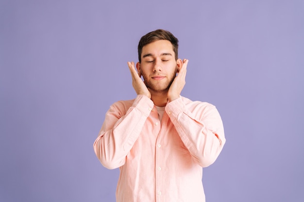 Portrait of calm young man covering ears with hands do not wanna listen standing on pink isolated background in studio with closed eyes. Concept of silence.