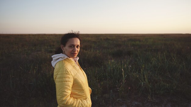 Photo portrait of calm young mixed race woman in yellow jacket looking at camera and standing against steppe surface at sunset