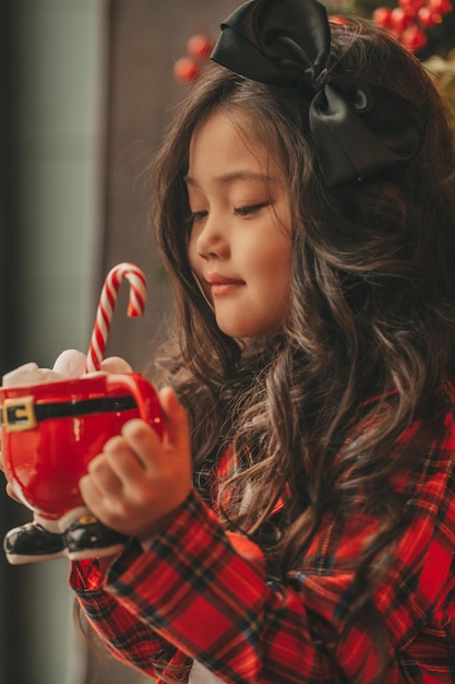 Foto ritratto di una bambina sorridente asiatica in pigiama a quadri rosso seduta con i regali a casa di natale