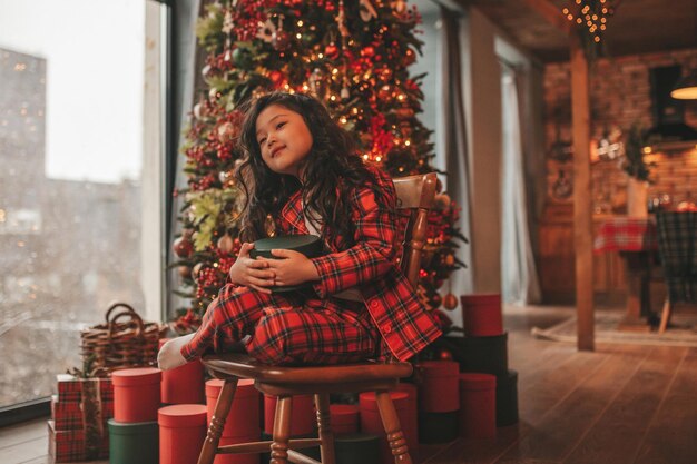 クリスマスの家でプレゼントを持って座っている赤の格子縞のパジャマで率直なアジアの笑顔の少女の肖像画