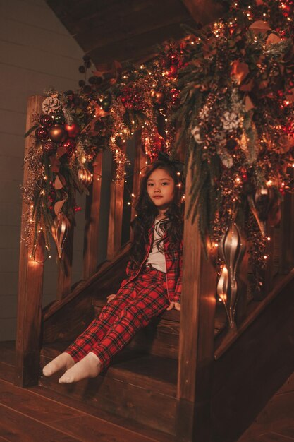 Portrait of candid asian smiling little girl in red plaid pajama sitting on wooden steps at Xmas