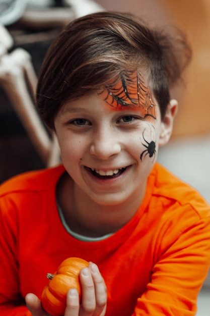 Portrait of candid authentic teen boy with face painted holds pumpkin at Halloween decorated party