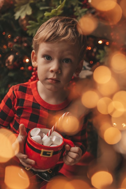 빨간색 격자 무늬 잠옷을 입은 솔직한 행복한 아이가 마시멜로와 사탕 지팡이가 있는 Xmas 머그를 들고 있습니다.