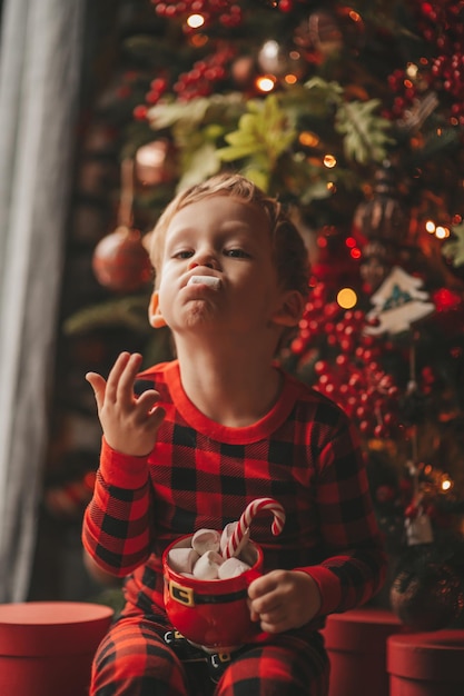 빨간색 격자 무늬 잠옷을 입은 솔직한 행복한 아이가 마시멜로와 사탕 지팡이가 있는 Xmas 머그를 들고 있습니다.