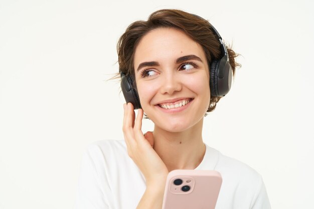 Photo portrait of candid young woman listens music in wireless headphones holding smartphone watching