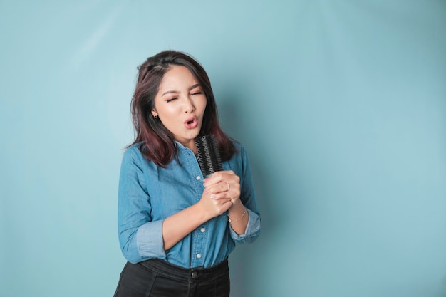 Portrait of carefree Asian woman having fun karaoke singing in microphone while standing over blue background