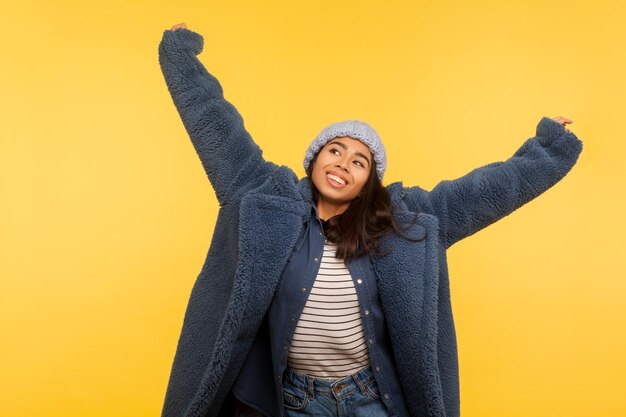 Portrait of carefree cheerful girl wearing warm winter hat and\
fur coat raising hands up smiling joyfully glamour fashion model in\
stylish urban outfit enjoying life indoor studio shot isolated