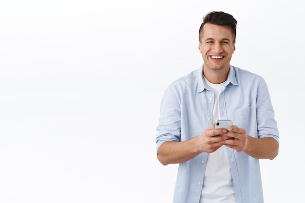 Portrait of carefree handsome caucasian man laughing over funny meme or blog post, holding mobile phone, texting friends on self-quarantine, covid19 lockdown, staying in touch online