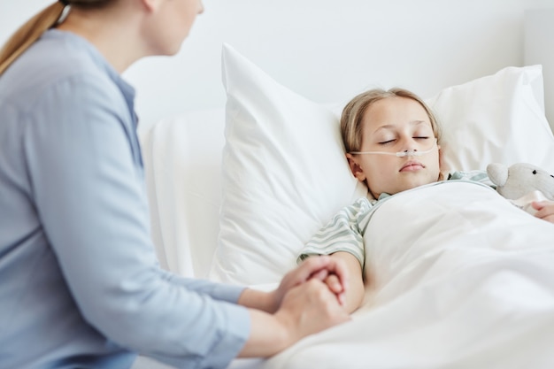 Portrait of caring mother holding hand of child in hospital room with oxygen support