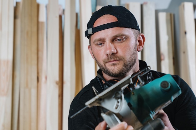 Portrait of a carpenter man in dark clothes holding a circular saw in his hands against the background of the workshop