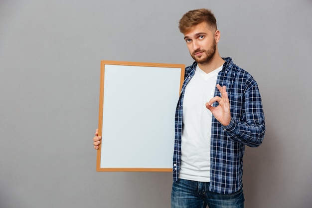 Portrait of a casual bearded man holding blank board and showing ok gesture isolated on a gray wall