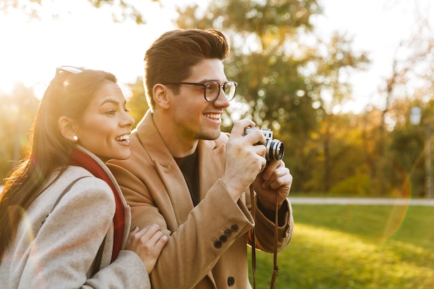 レトロなカメラで写真を撮って、秋の公園を歩きながら笑っているカジュアルなカップルの男女20代の肖像画