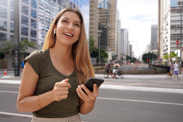 Портрет случайной девушки с беспроводными наушниками, смотрящей вверх и использующей телефон на городском проспекте в летнее время с размытыми людьми на заднем плане
