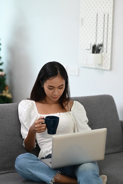 Portrait of casual woman using laptop and drinking coffee while sitting on couch at home.