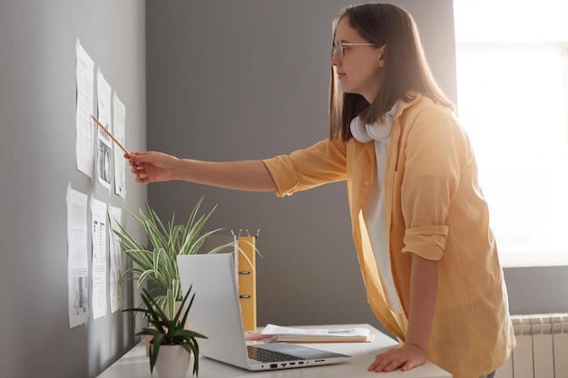 Portrait of Caucasian beautiful woman with brown hair wearing yellow shirt posing in office standing near her workplace and checking information on graphs on wall looking with serious face