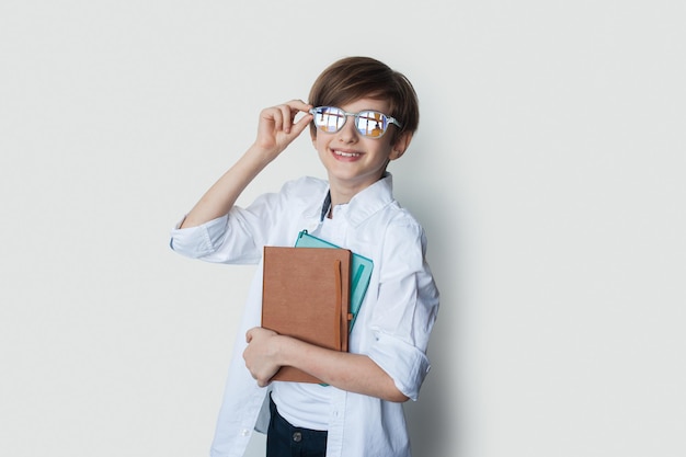 Portrait of a caucasian boy in white shirt holding a pile of copy books who stands smiling and holds...