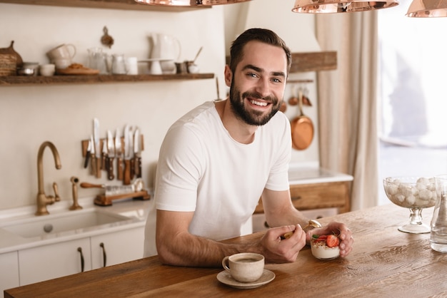 Portrait of caucasian brunette man  eating panna cotta dessert with teaspoon while having breakfast in stylish kitchen at home