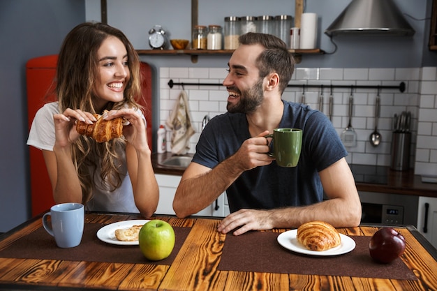 Portrait of caucasian couple man and woman eating croissants while having breakfast in stylish kitchen