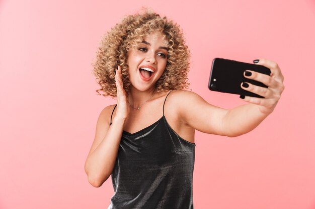 Portrait of caucasian curly woman 20s smiling and taking selfie photo on smartphone