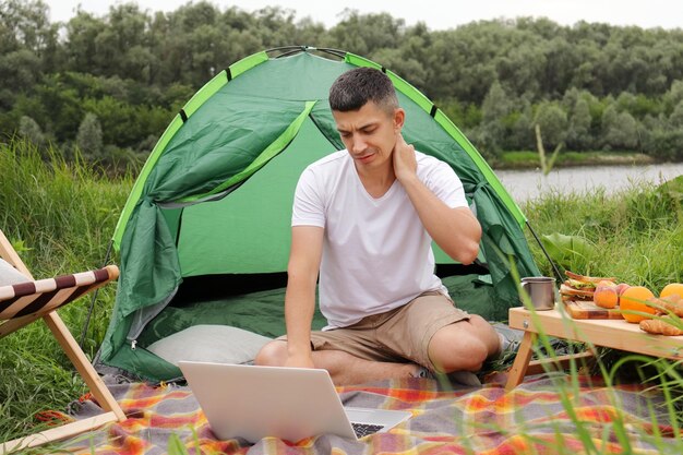 Ritratto di libero professionista caucasico uomo dai capelli scuri seduto all'aperto su plaid a terra che lavora online su laptop essendo stanco ed esausto sente dolore al collo guardando lo schermo del notebook