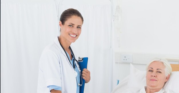 Portrait of caucasian female doctor holding clipboard smiling while female patient in hospital bed