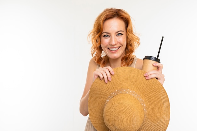 Portrait of caucasian female with fair red hair, pretty face in beautiful light dress and big hat smiles and drinks coffee