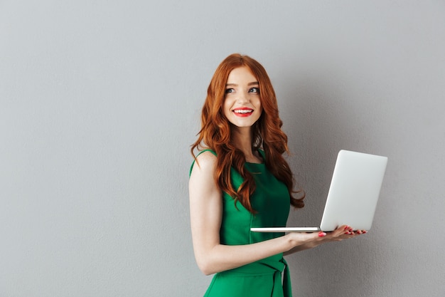 Portrait of caucasian ginger long haired woman in green dress looking aside and holding white laptop in hands, isolated over gray wall