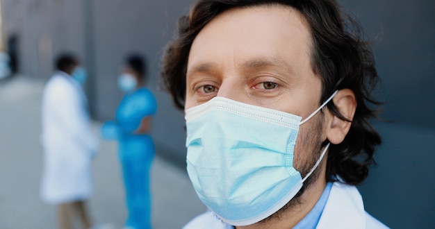 カメラを見ている医療マスクの白人ハンサムな男の医者の肖像画。呼吸保護の男性医師のクローズアップ。背景にアフリカ系アメリカ人の医者の同僚。