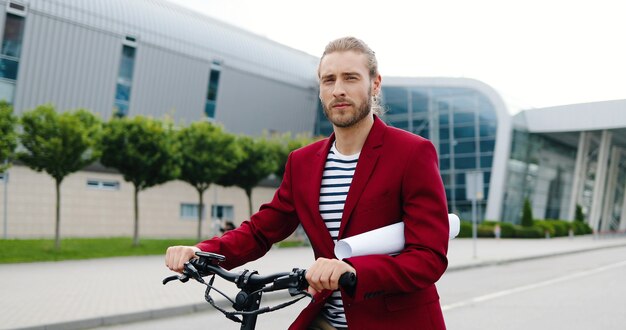 バイクや電動スクーターで屋外に立って、カメラを見ている赤いジャケットの白人ハンサムな若い男の肖像画。紙を持っているか、街の通りに絵を描く自転車でかっこいいスタイリッシュな男