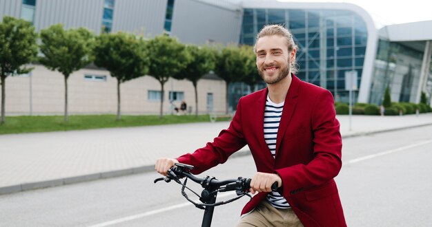 Portrait of Caucasian handsome young man in red jacket standing outdoor with bike or electric scooter and smiling to camera. Good-looking stylish guy at bicycle on city street.