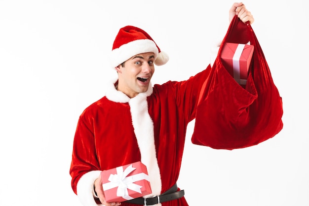 Portrait of caucasian man 30s in santa claus costume and red hat holding festive bag with gift boxes