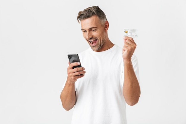 白で隔離のスマートフォンとプラスチックのクレジットカードを保持しているカジュアルなTシャツを着ている白人男性30代の肖像画
