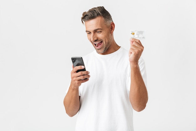 Portrait of caucasian man 30s wearing casual t-shirt holding smartphone and plastic credit card isolated on white