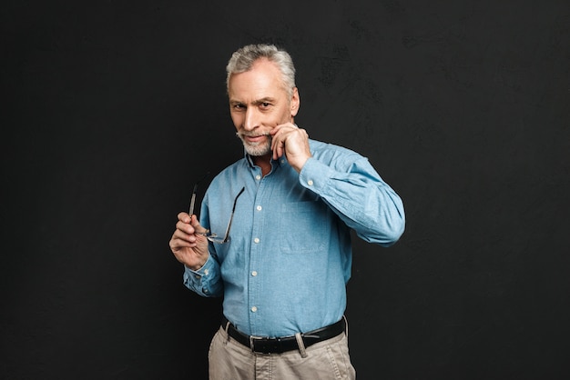 Portrait of caucasian man 60s with grey hair and beard holding glasses while touching his gray mustache, isolated over black wall