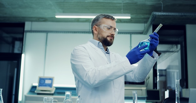 Foto ritratto dell'uomo caucasico in vetri e abito bianco che fa un'analisi del liquido blu nella provetta durante la ricerca medica o farmaceutica in laboratorio.