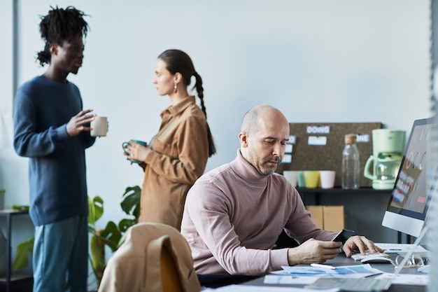 Portrait of caucasian mature man using laptop at workplace with\
people chatting in background in com