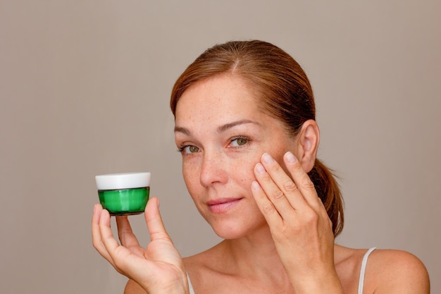 Portrait of caucasian middle aged woman face with freckles showing soft skin by fingers holding cream on white background