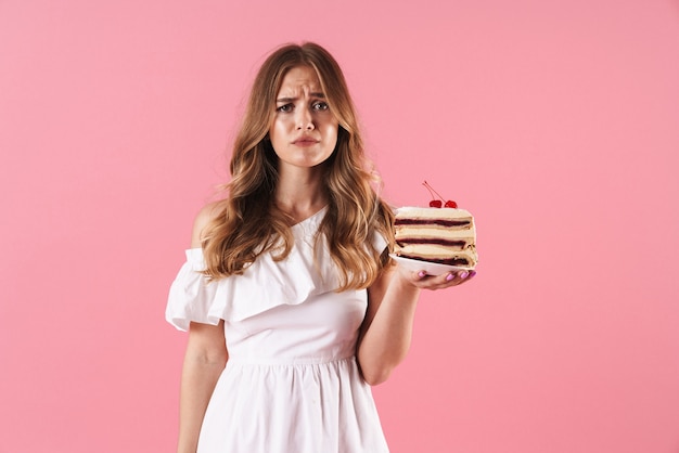 Portrait of caucasian sad woman wearing white dress looking at front and holding piece of cake isolated over pink wall