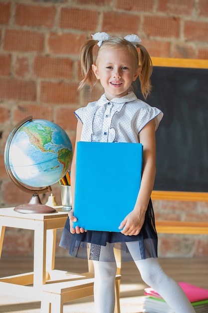 Portrait of caucasian schoolgirl standing in front of chalkboard with book in her hands, back to school concept