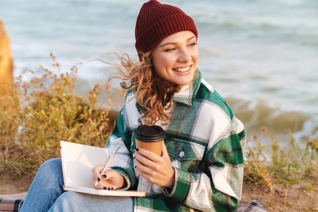 Portrait of caucasian smiling woman drinking coffee takeaway and making notes in diary while sitting on blanket by seaside