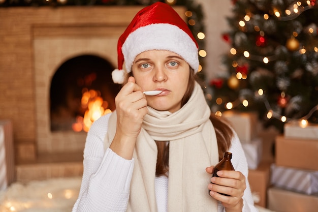 Portrait of Caucasian unhealthy woman wearing santa claus hat, being wrapped in warm scarf, sitting near Christmas tree. drinking syrup, suffering from cough, catching cold during winter holidays.