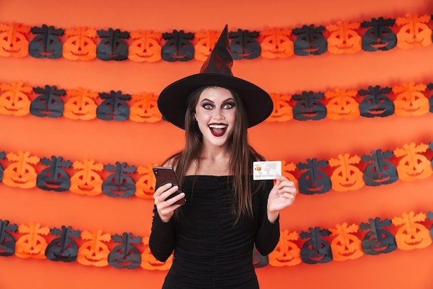 Foto ritratto di una strega caucasica in costume nero di halloween che tiene in mano smartphone e carta di credito isolata sulla parete di zucca arancione