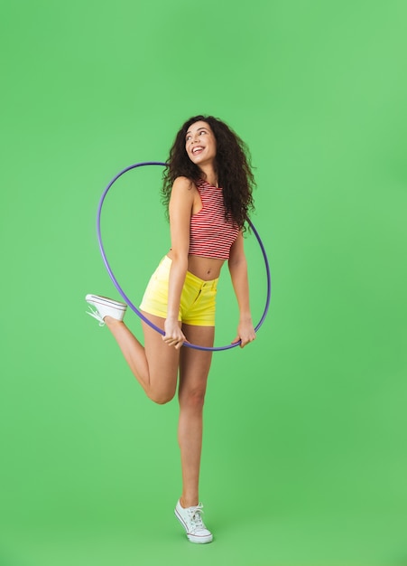 portrait of caucasian woman 20s wearing summer clothes doing exercises with hula hoop during workout on green wall