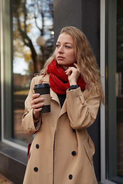 Portrait of caucasian woman in beige trench coat outdoors with\
cup of coffee