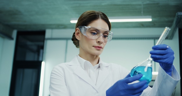 Portrait of the Caucasian woman in glasses and white robe doing an analysis of the blue liquid in the test tube during medical or pharmaceutical research and in the laboratory.