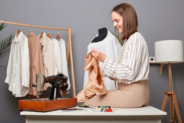 Portrait of caucasian woman in sewing workshop holding detail of new dress sitting on table smiling designer creating new collection