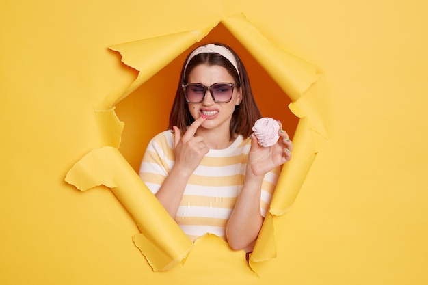 Portrait of Caucasian woman wearing hair band and striped t shirt felling discomfort in her teeth pointing holding marshmallow standing in yellow paper hole